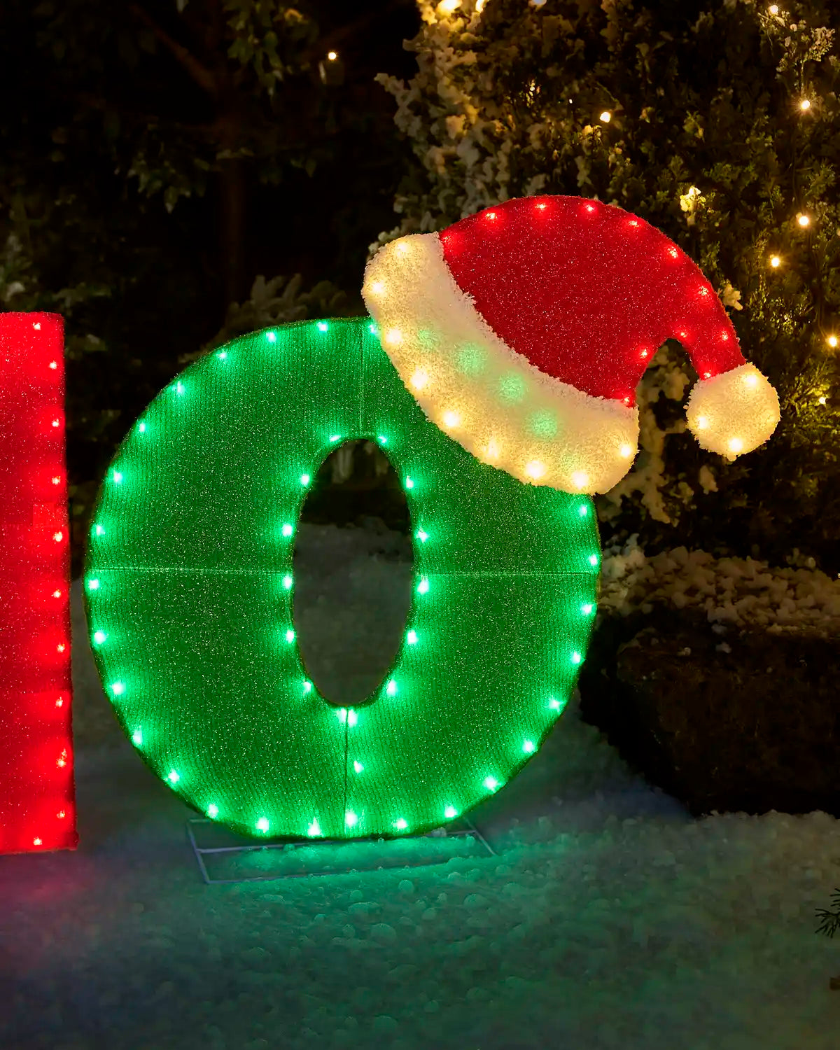 A large illuminated Christmas decoration shaped like the number 0 in bright green, with a red and white Santa hat on top, displayed outdoors in snow at night.