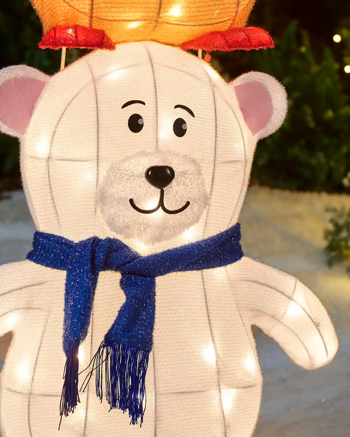 A white polar bear-shaped decorative lantern with warm white string lights, featuring a blue glittery scarf and a glowing orange lantern on its head, photographed outdoors at night.
