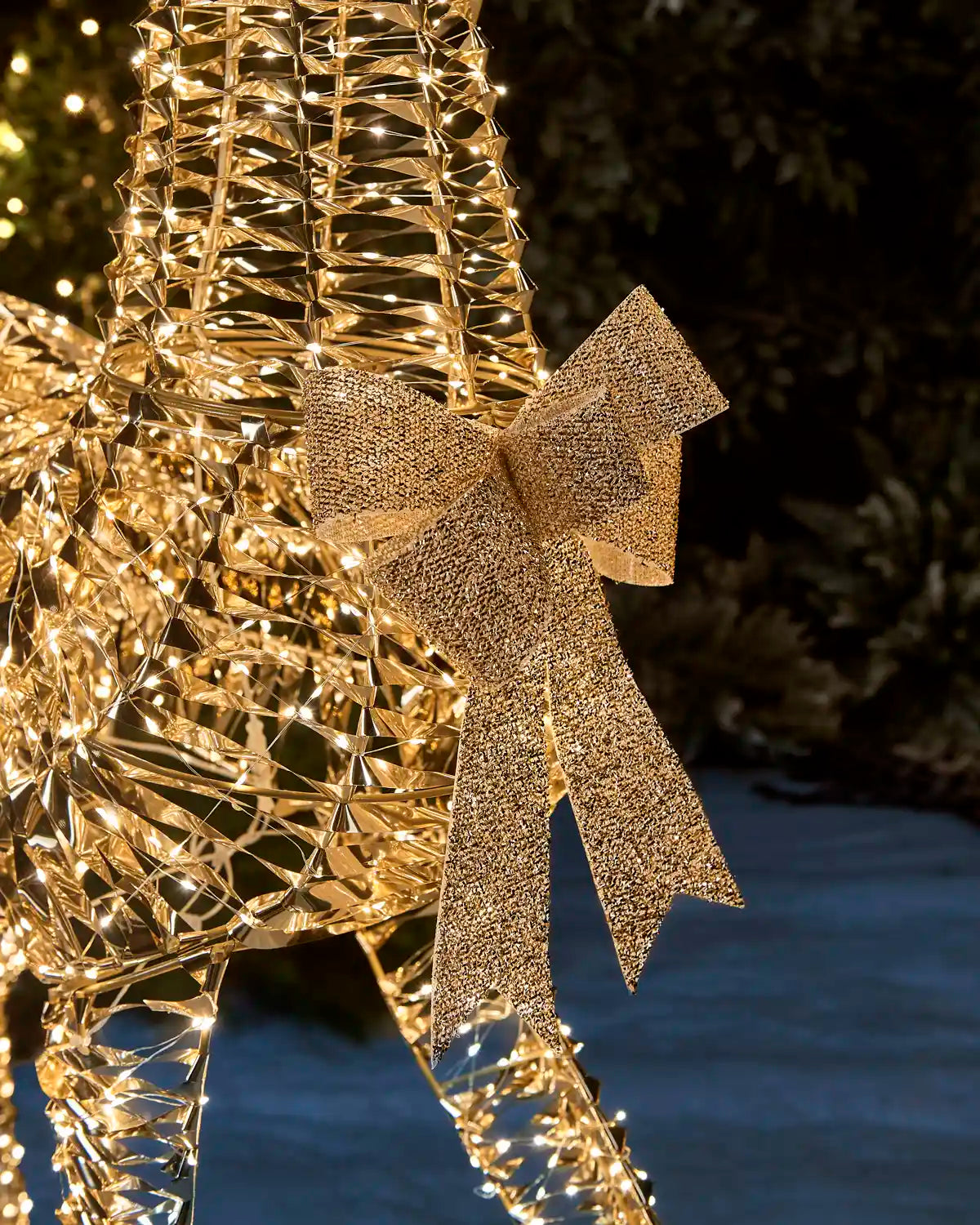 A close-up of a large gold wire holiday decoration, part of a reindeer or tree display, adorned with a glittery gold bow and glowing warm white string lights, set against a dark outdoor background.