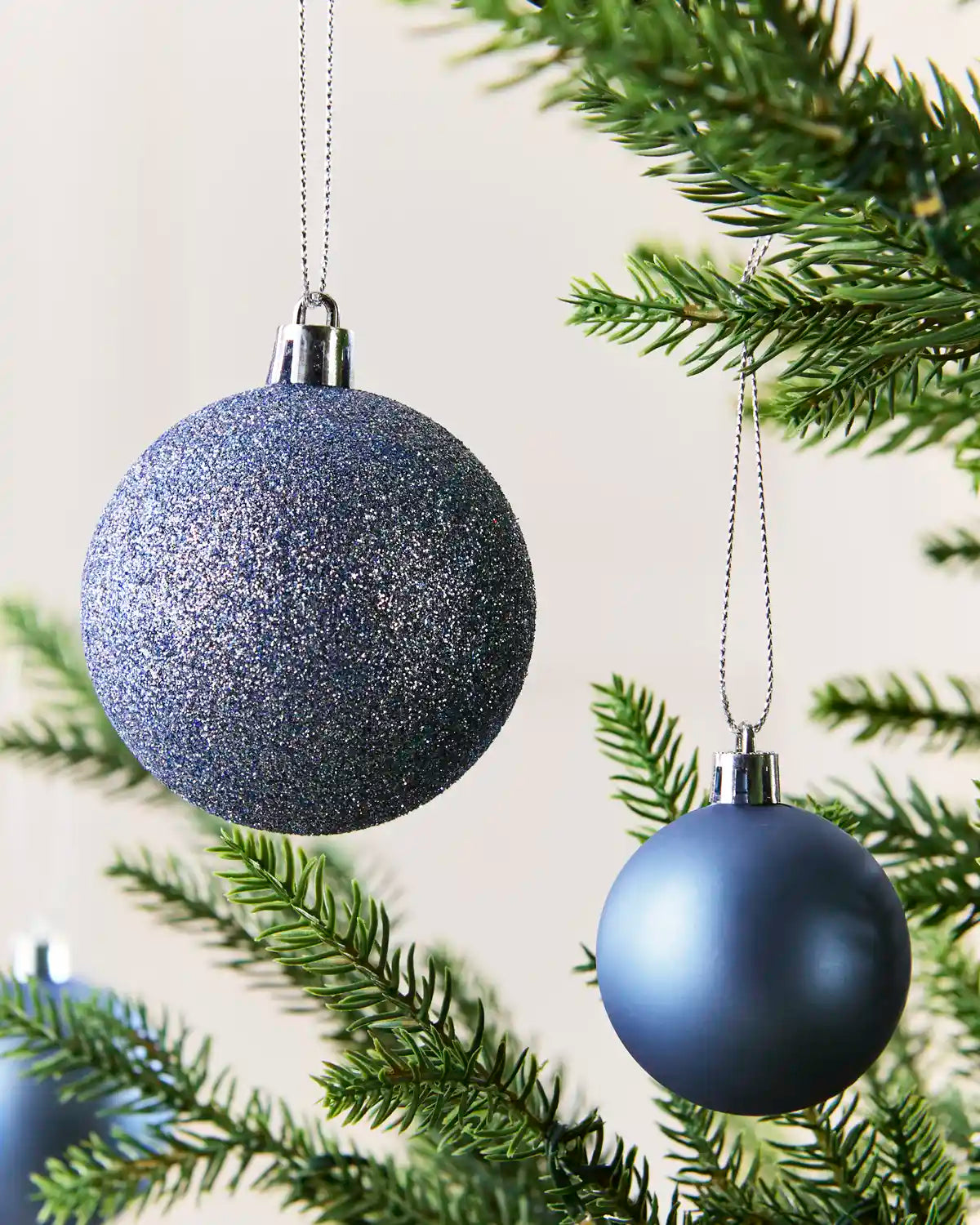 Two Ice blue Christmas tree ornaments hanging from green pine branches against a soft cream background.