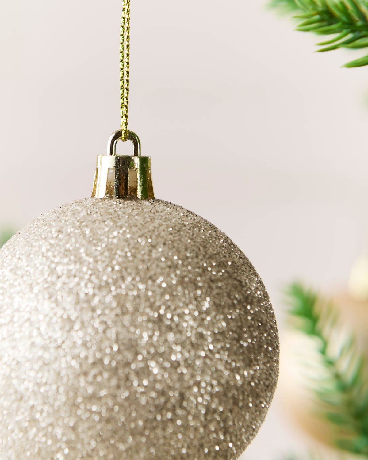 A close-up of a glittery Latte Christmas ornament hanging from a gold chain, with soft blurred green pine branches in the background.