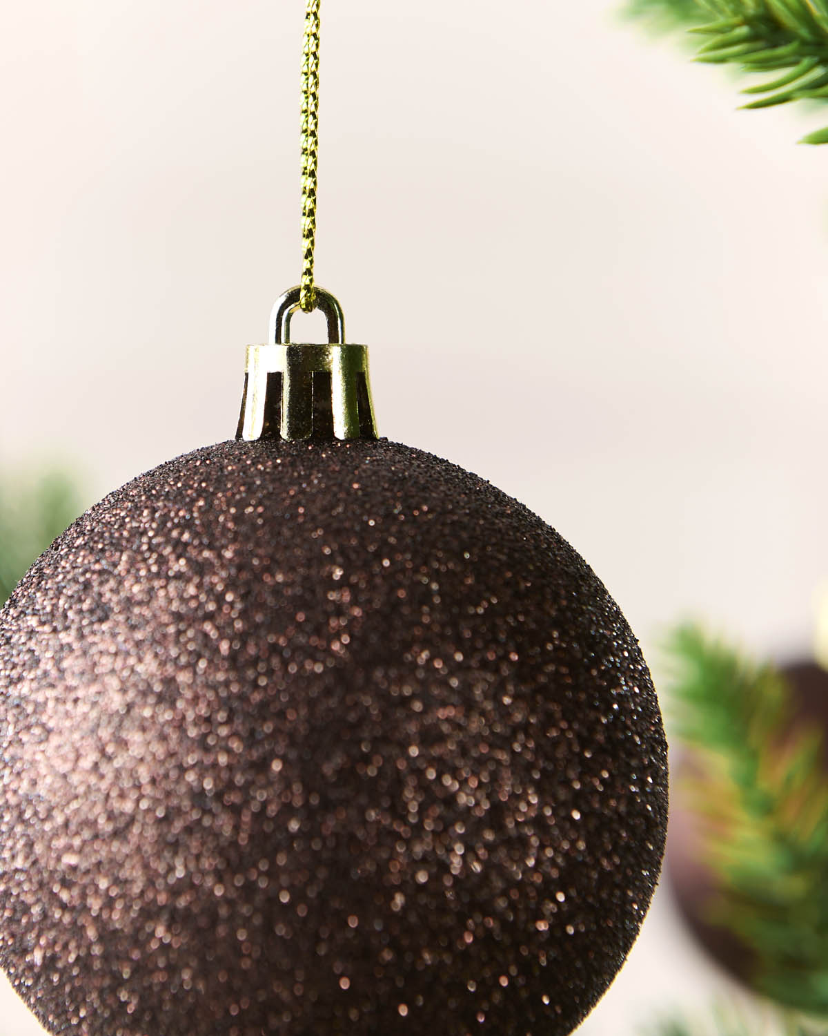 A close-up of a glittery Espresso Christmas ornament hanging from a gold chain, with blurred green pine branches in the background.