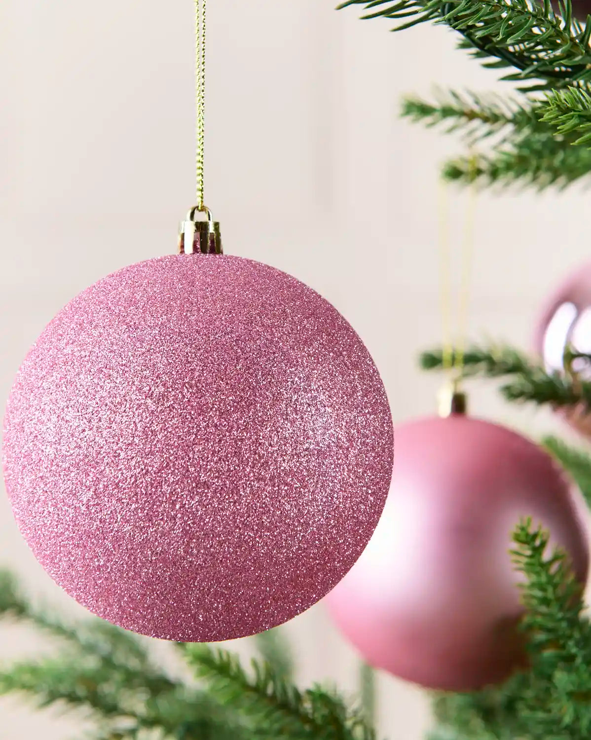 A close-up of a glittery pink Christmas bauble hanging from a gold string on a Christmas tree, with another matte pink bauble blurred in the background.