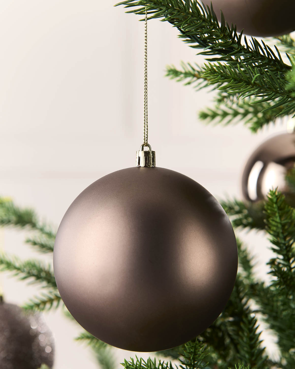 A matte mocha Christmas bauble ornament hanging from a gold string on a green Christmas tree branch.