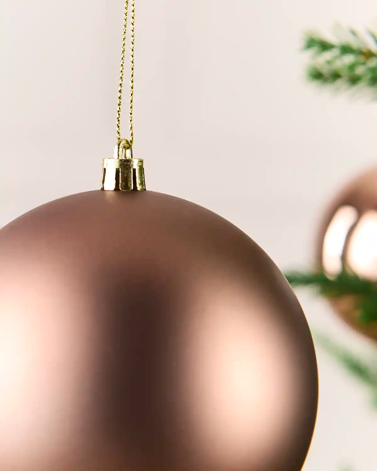 A close-up of a matte metallic mocha Christmas ornament hanging from a gold string, with a softly blurred background showing green pine branches and another ornament.