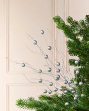Silver berry branch garland with small metallic silver spheres on thin wire stems, shown draped over green pine tree branches against a cream paneled wall.