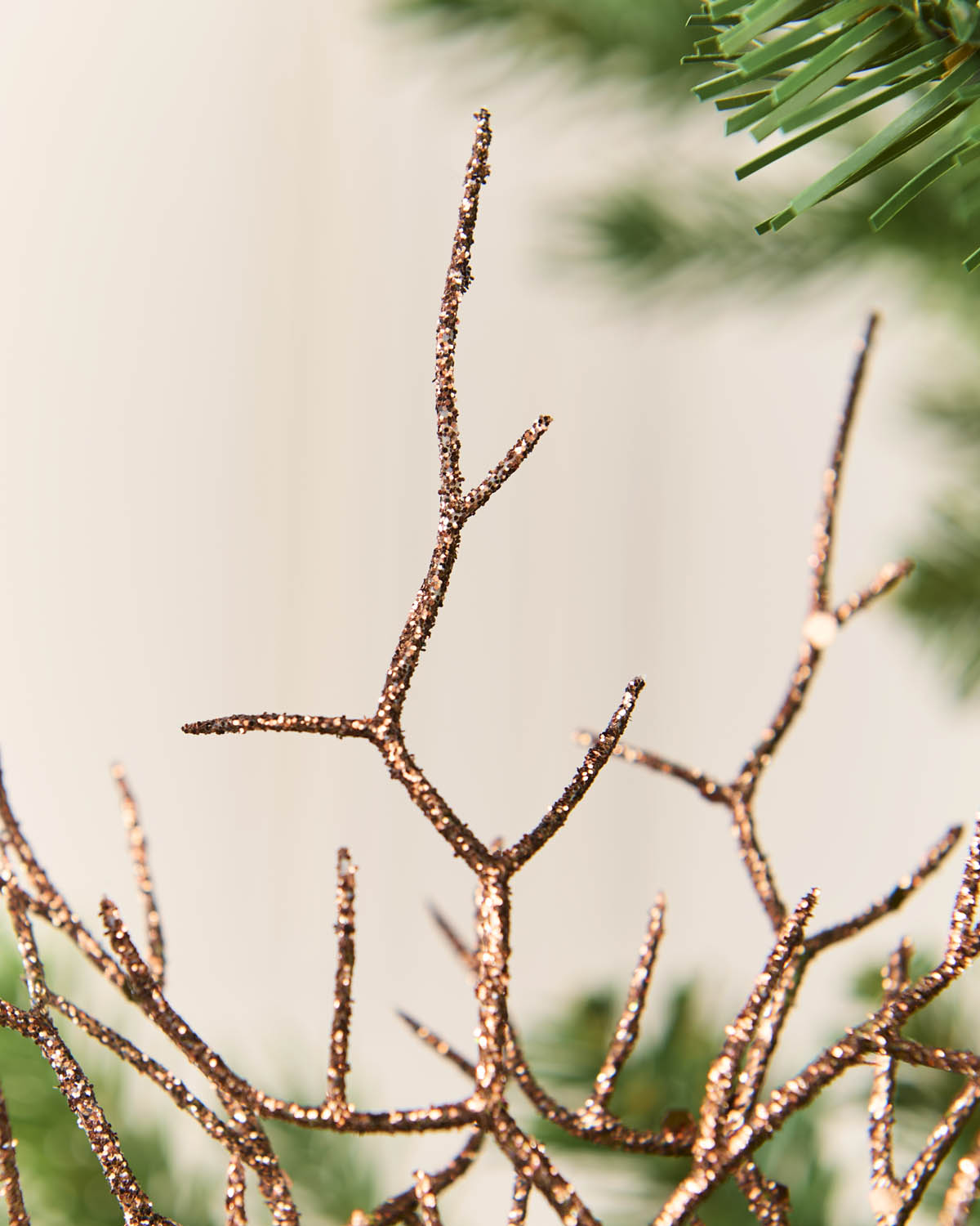 Mocha glittery twig tree branch holiday decoration with a sparkling finish, shown in a close-up shot against a soft beige background with blurred green pine needles.
