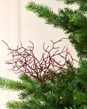 A decorative coral ornament with a glittery burgundy red finish, styled among green pine needles against a soft cream background.