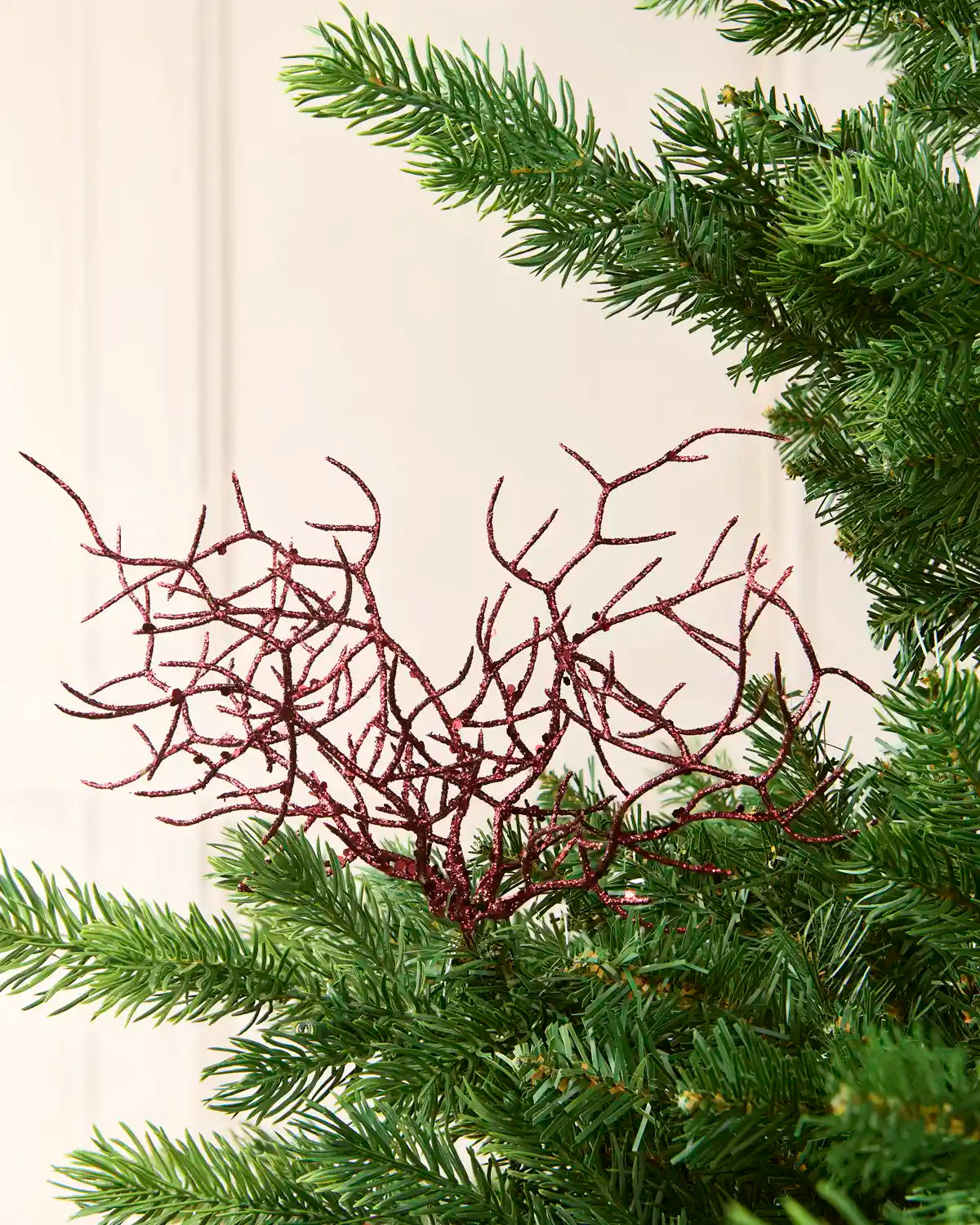 A decorative coral ornament with a glittery burgundy red finish, styled among green pine needles against a soft cream background.