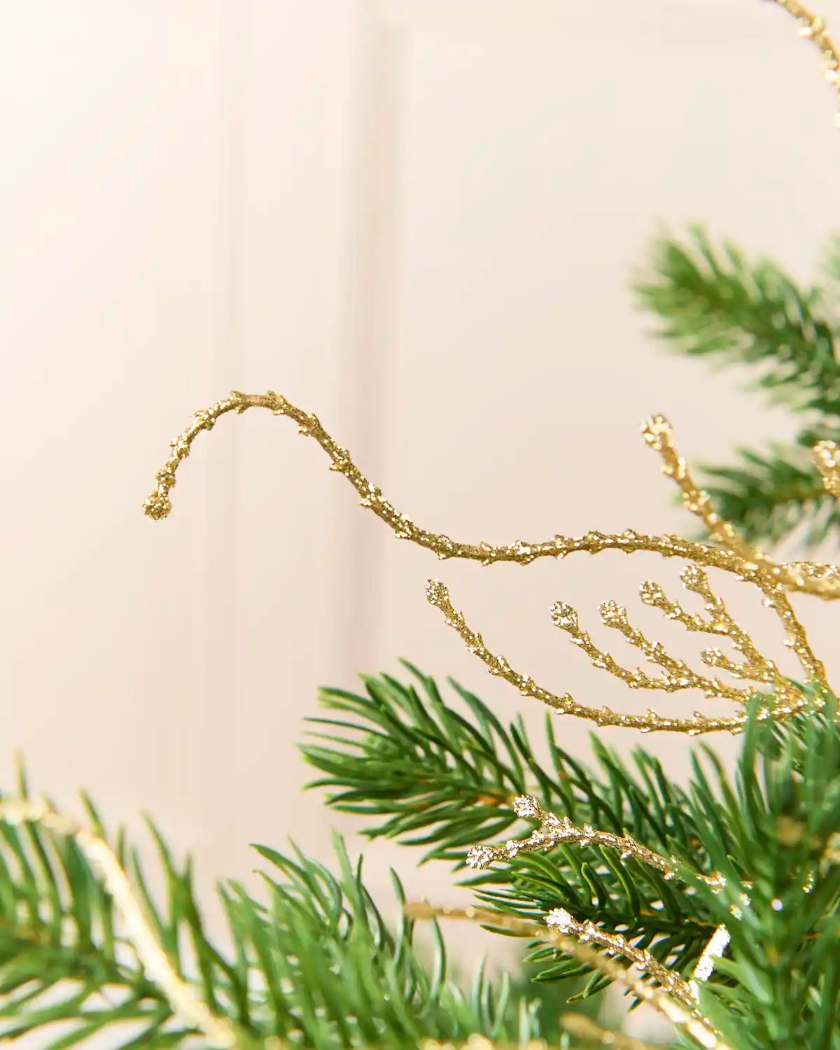 Close-up of green pine branches decorated with glittery gold tinsel garlands against a soft cream background.