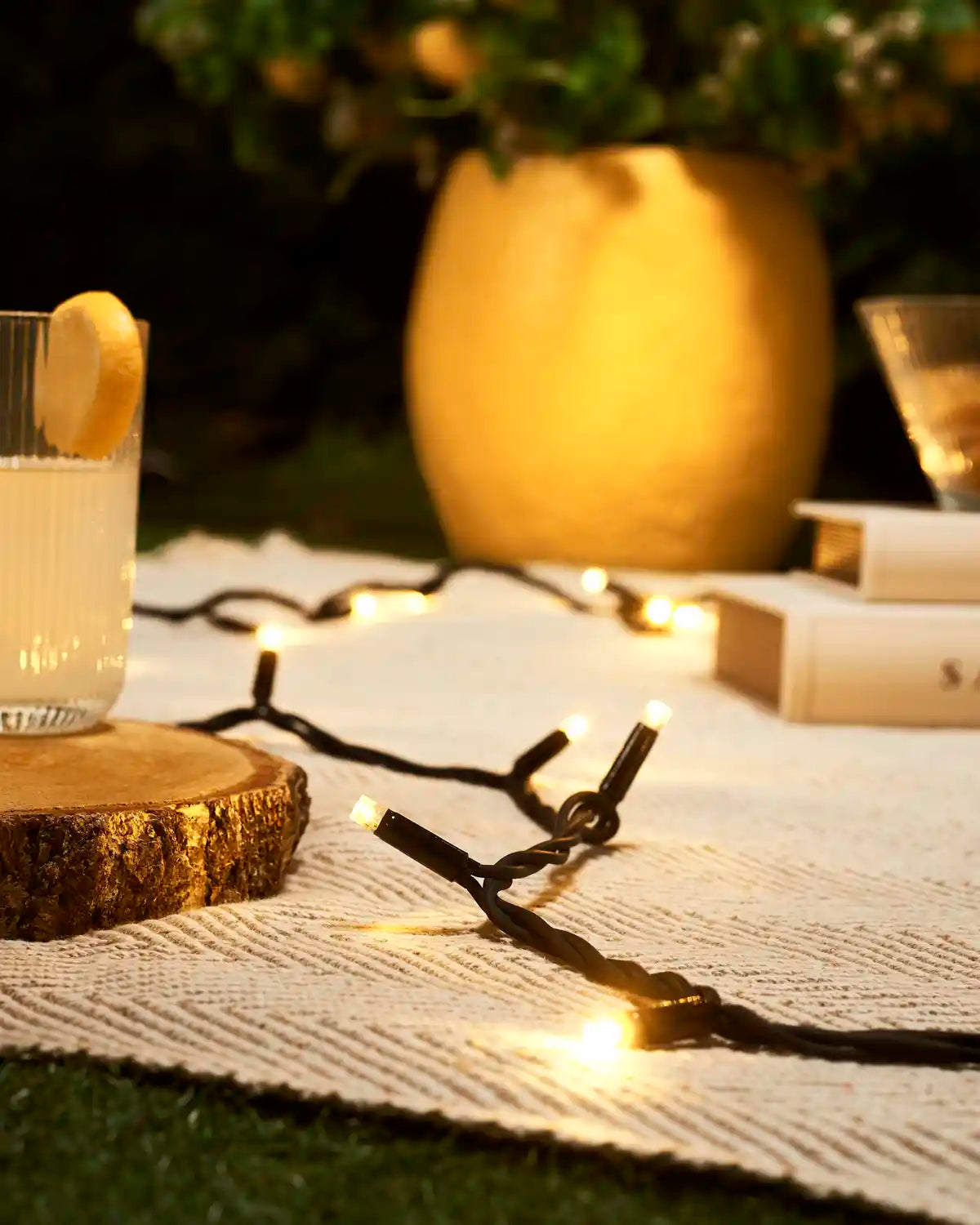 Warm White LED string light on white cloth with blurry yellow plant pot in the background, blurry book stack and wooden coaster with a drink on. 