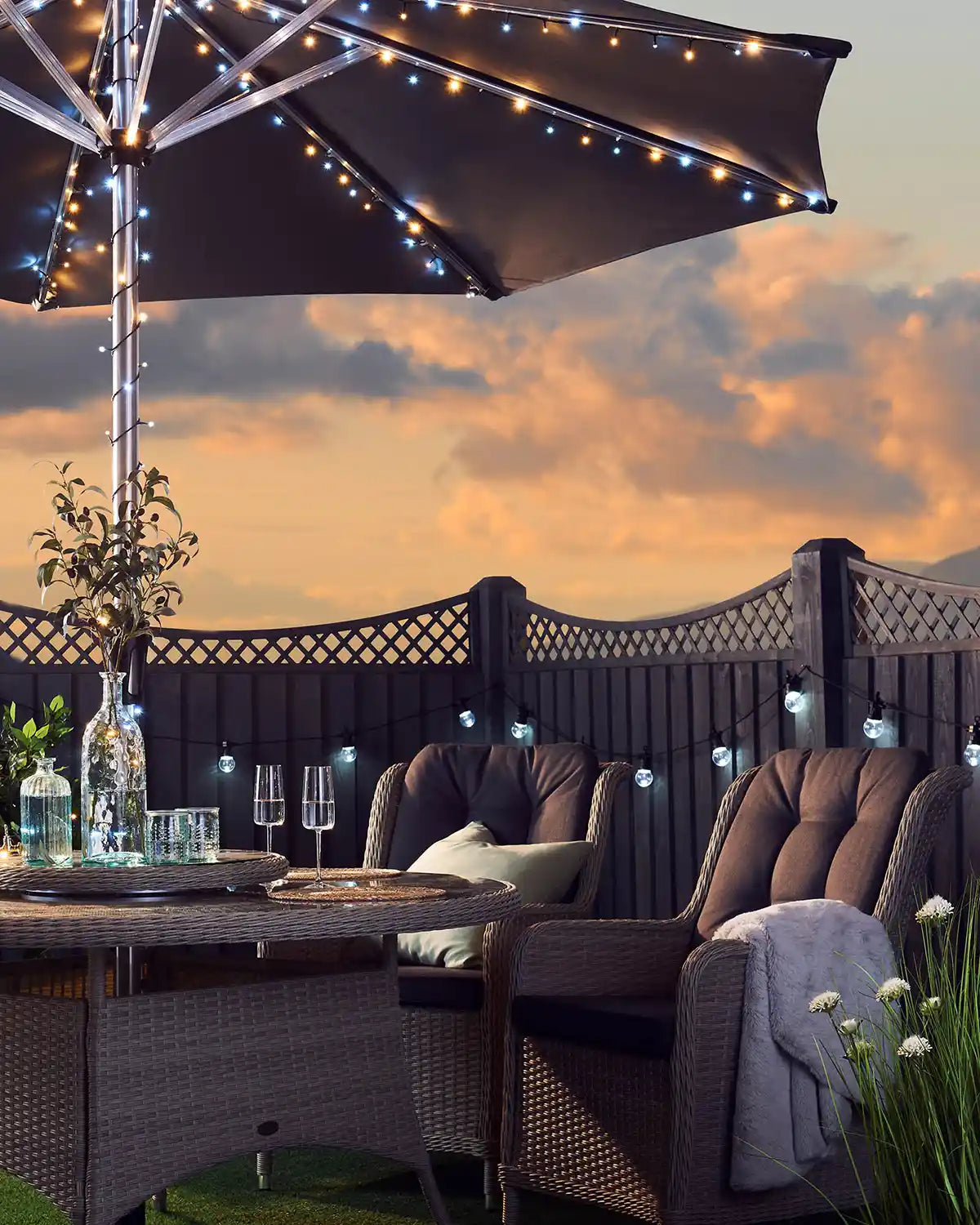 Warm white and ice white sting lights wrapped around an outdoor umbrella with a sun set background and outdoor furniture