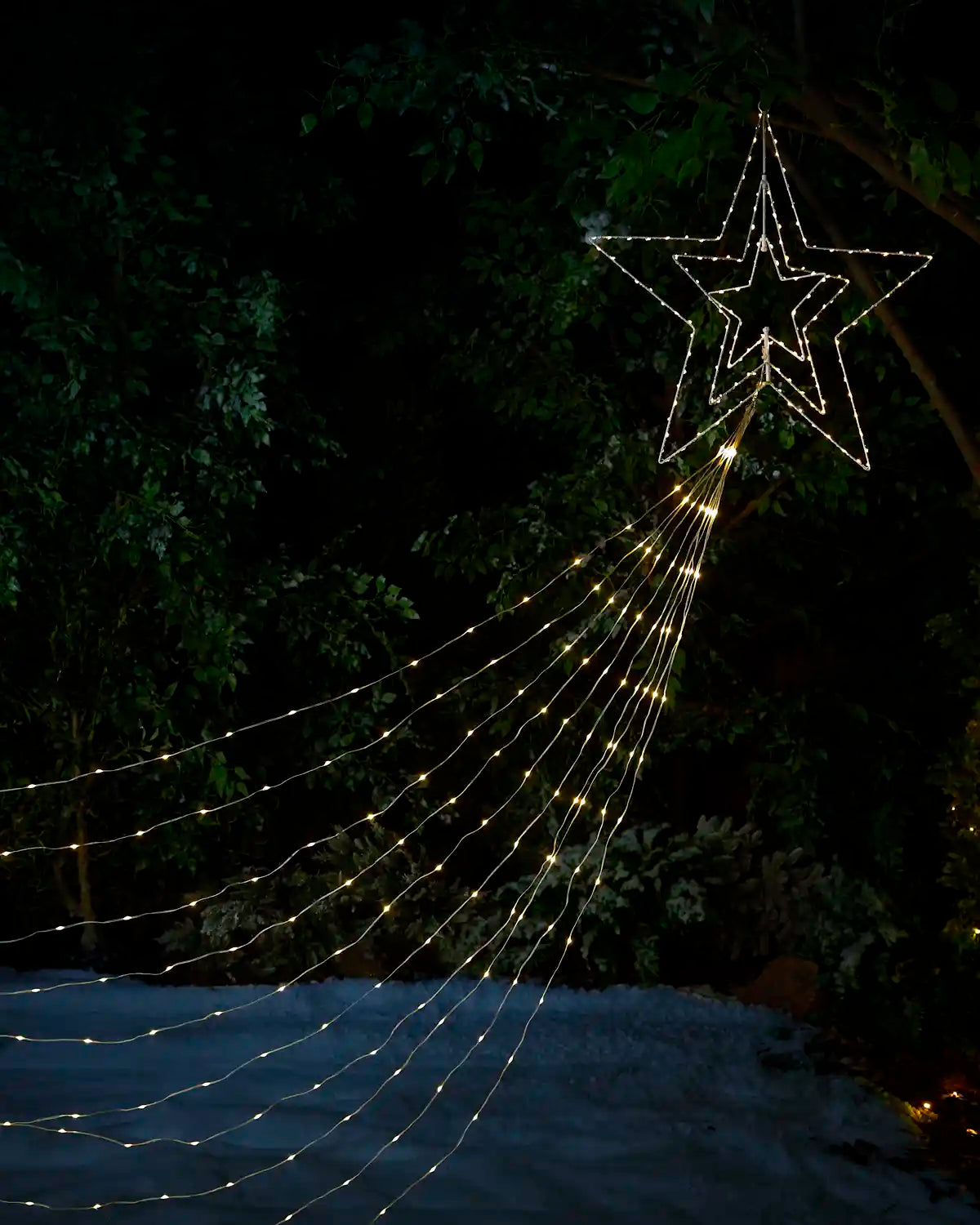 Warm white string lights shaped like a large star with trailing lines, displayed outdoors at night against dark trees and snow.