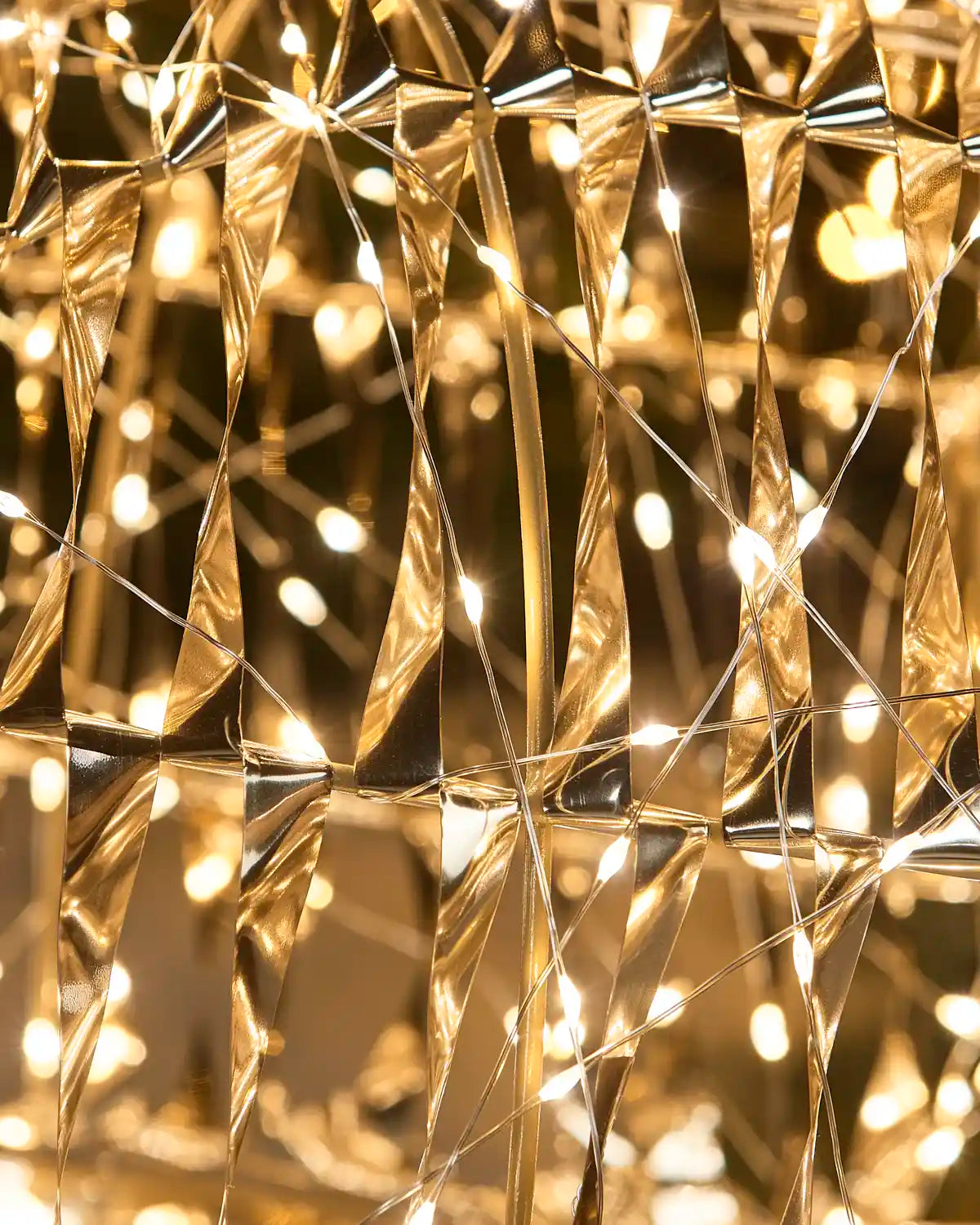 Warm white string lights woven through a faceted, diamond-like metallic mesh structure, creating a glowing golden and bronze pattern with soft bokeh light spots.