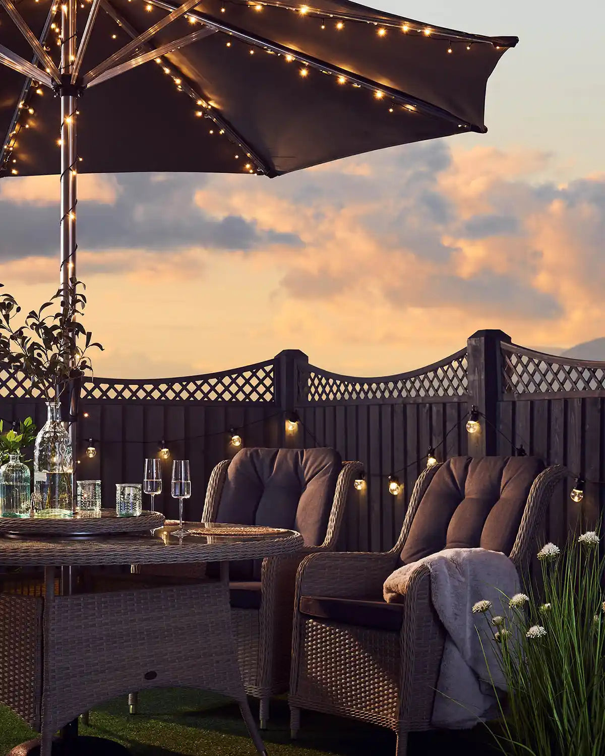 Warm white string lights wrapped around an outdoor umbrella with outdoor furniture against a sun set background 