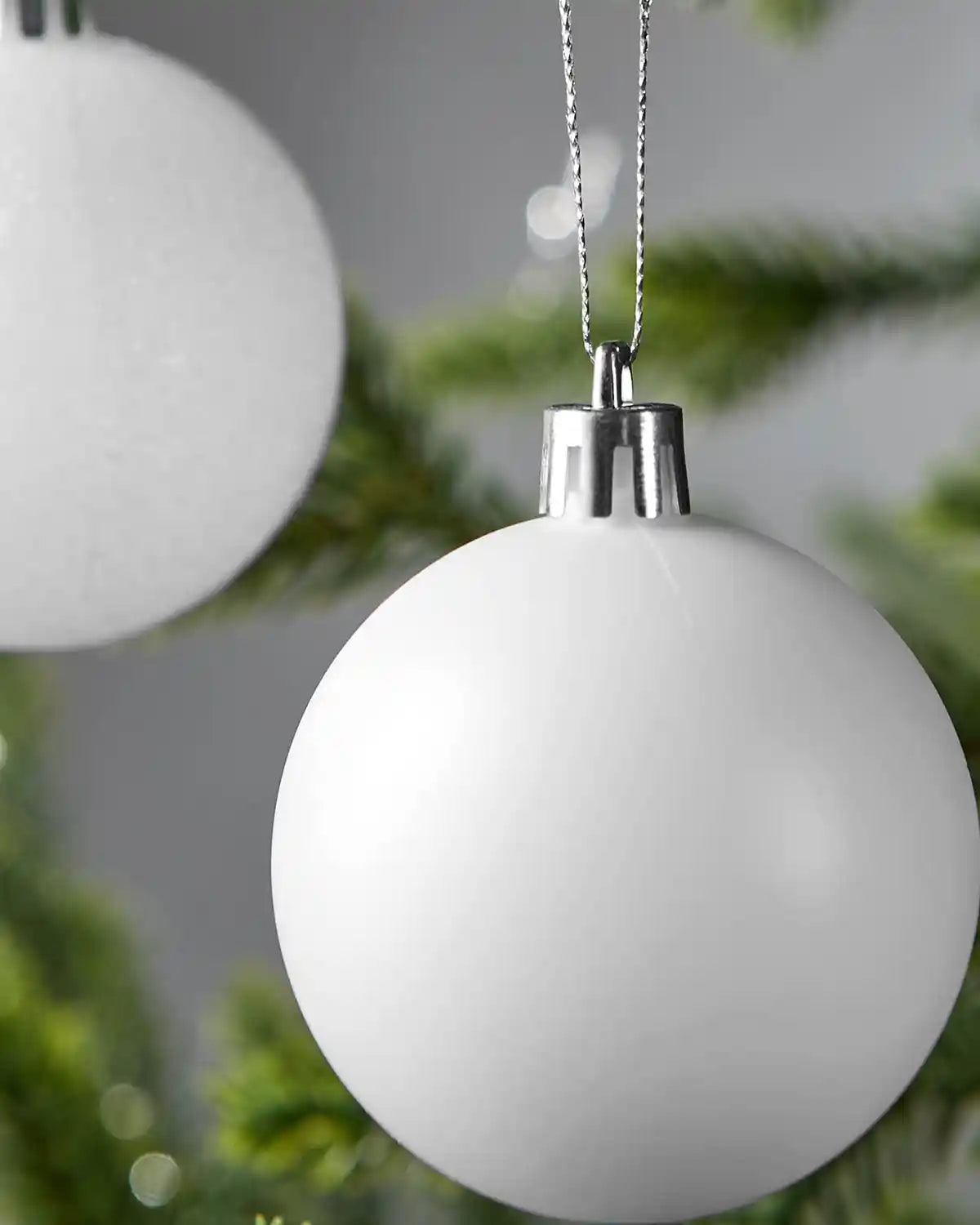 White Christmas bauble on a Christmas tree branch with a blurry bauble in the background against a Grey wall