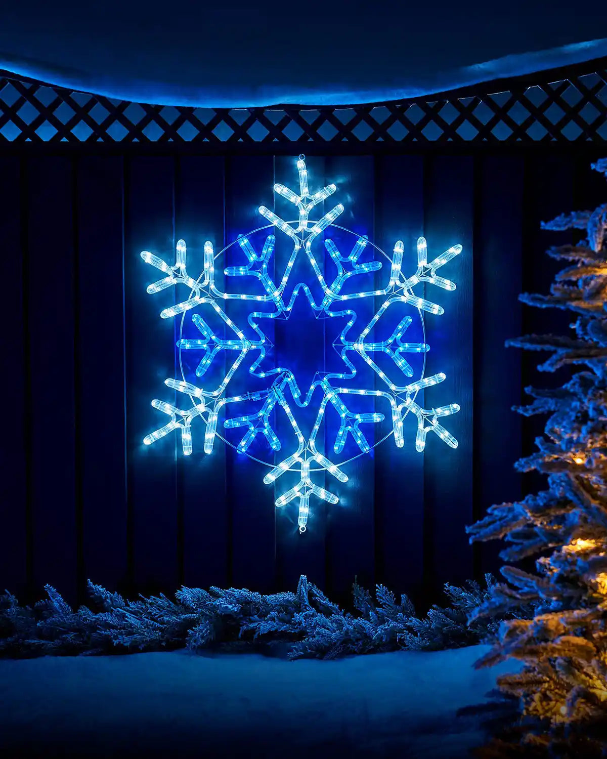 White and Blue LED lights in the shape of a Christmas Snowflake on a garden fence with a snowy background and a Christmas tree