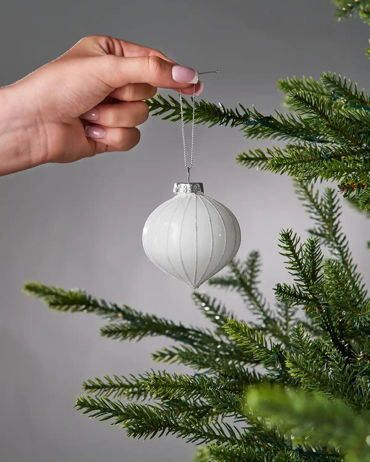 White bauble being hung on a Christmas tree branch against a grey background