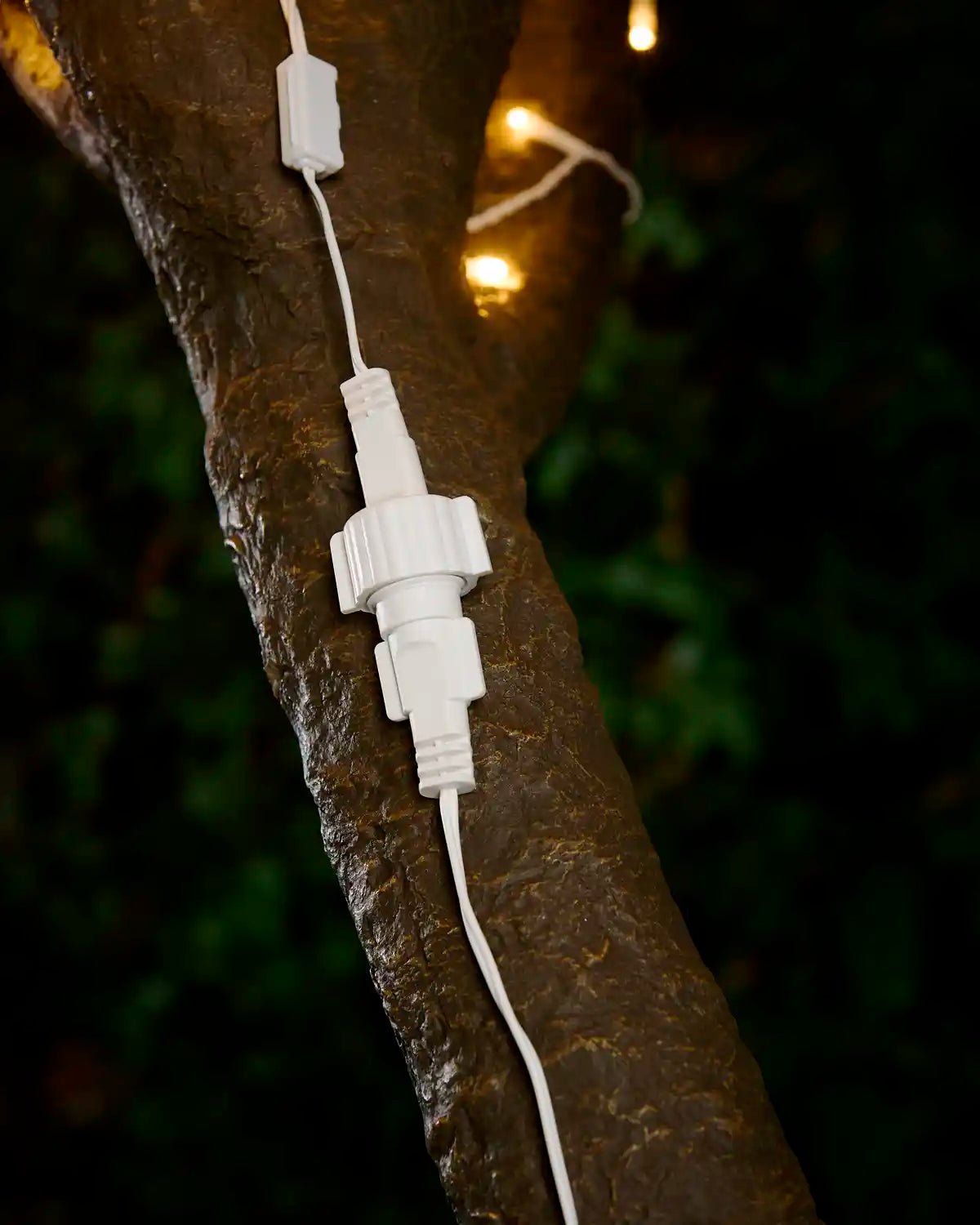 White connection point on extension cable with screw cap attached to tree with blurry warm white LEDs in the background