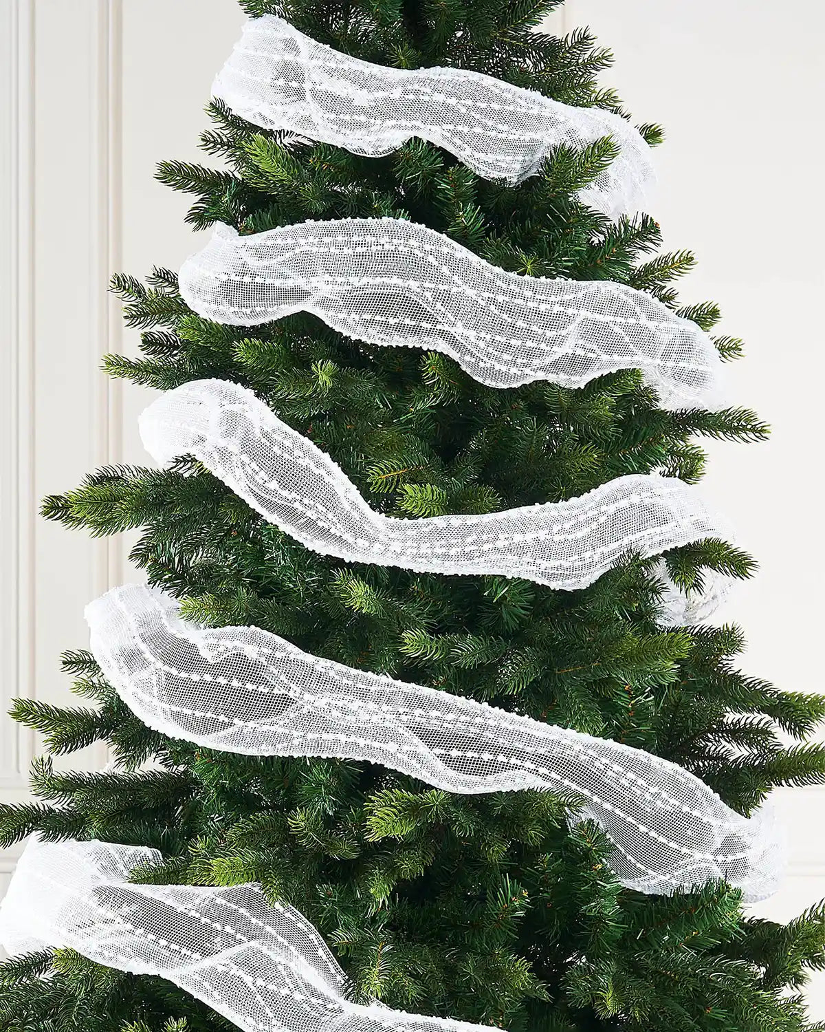 White decorative mesh ribbon with lace pattern wrapped around Christmas tree against cream wall