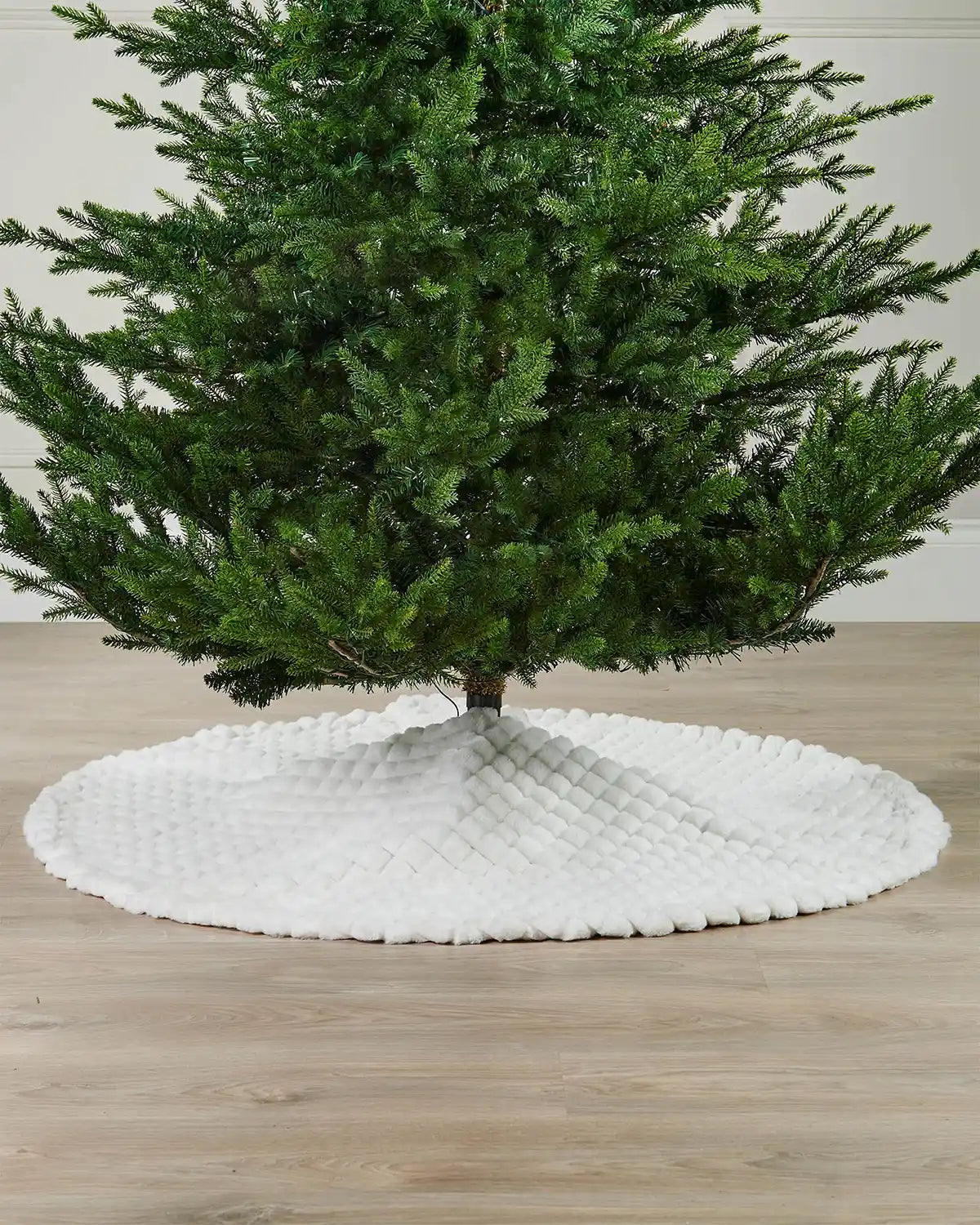 White diamond faux fur circular tree skirt on light wooden flooring underneath the base of a green Christmas tree