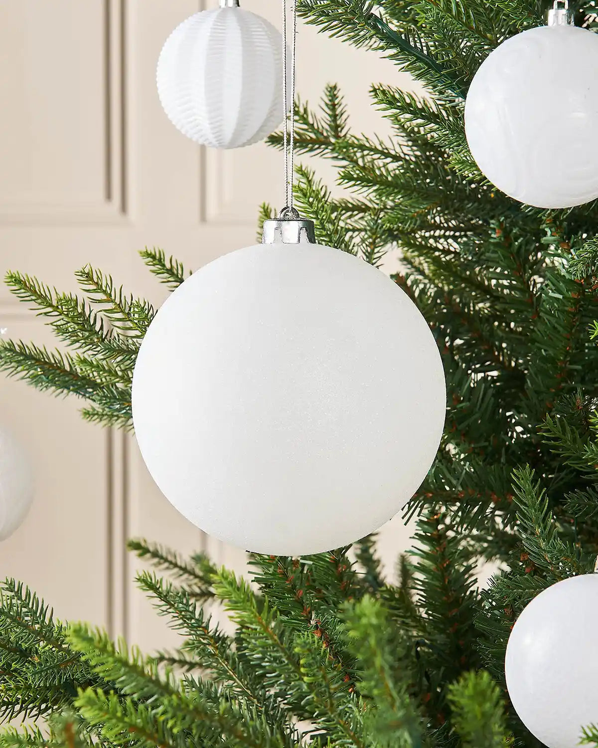 White glitter oversized 15cm bauble on a Christmas tree branch with 4 smaller white baubles in the background