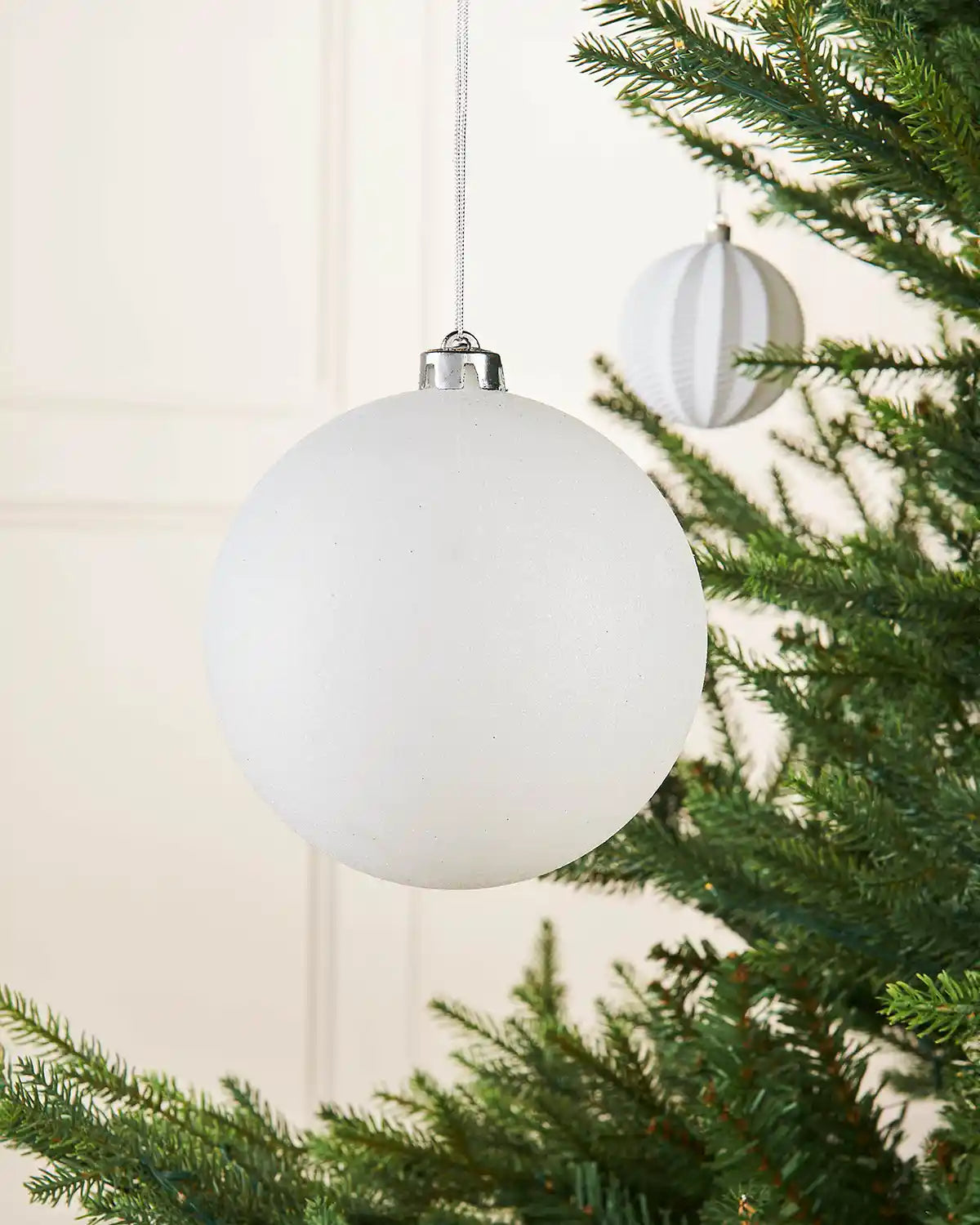 White glitter oversized 15cm bauble on a Christmas tree branch with a smaller white bauble in the background