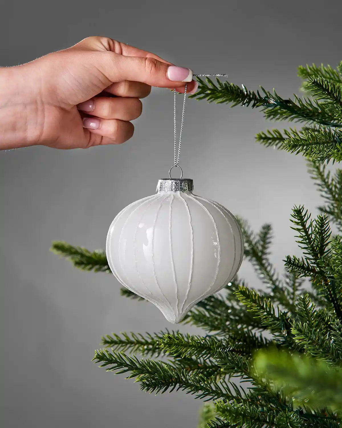 White ornament being hung on a Christmas tree branch against a gray background