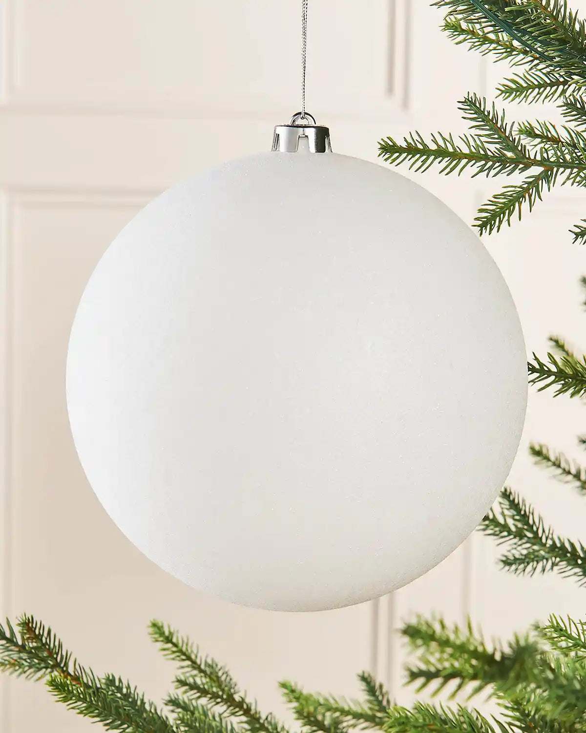 White oversized glitter bauble on a Christmas tree branch against a cream panel wall