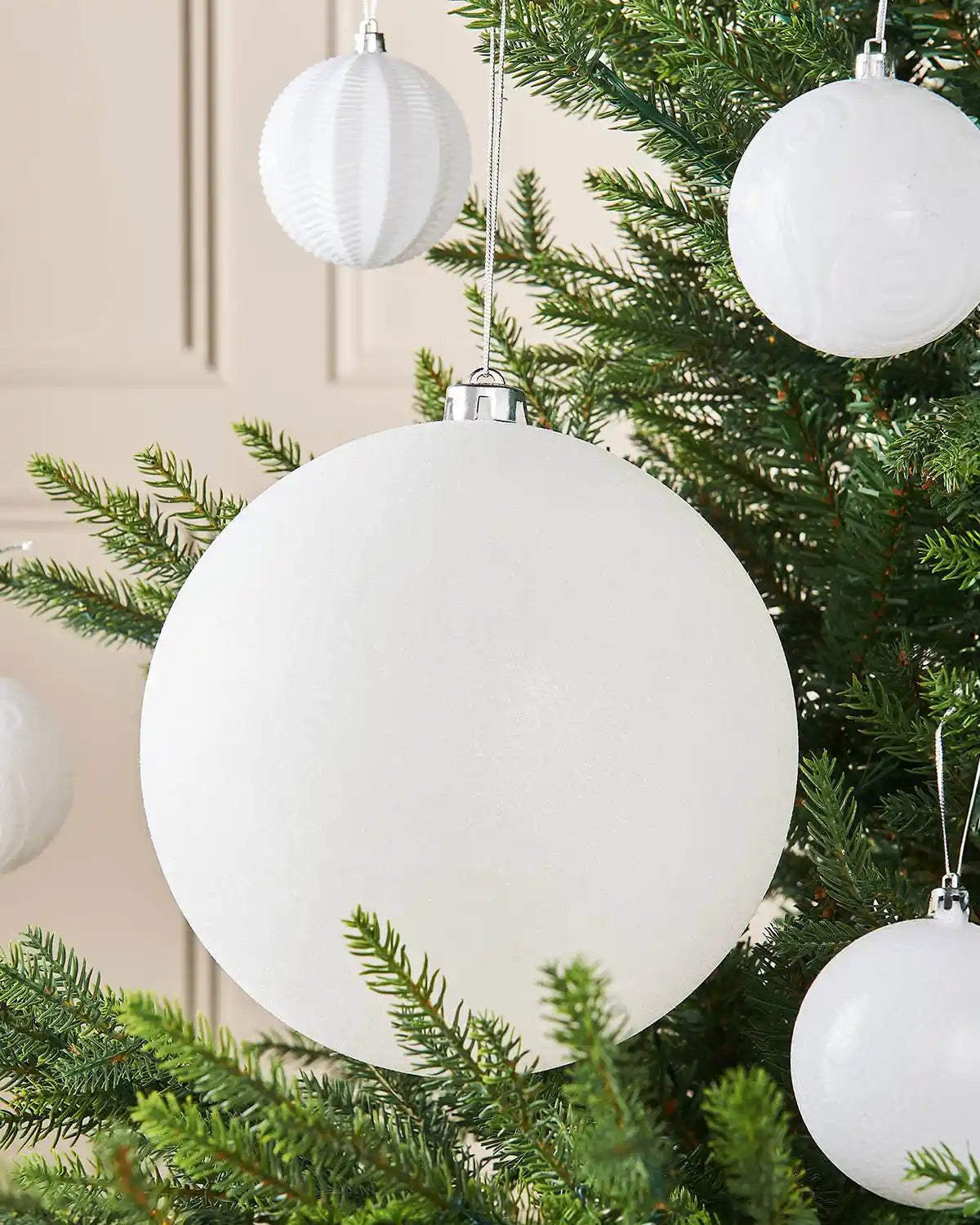 White oversized glitter bauble on a Christmas tree branch against a cream panel wall with 4 smaller white baubles in the background