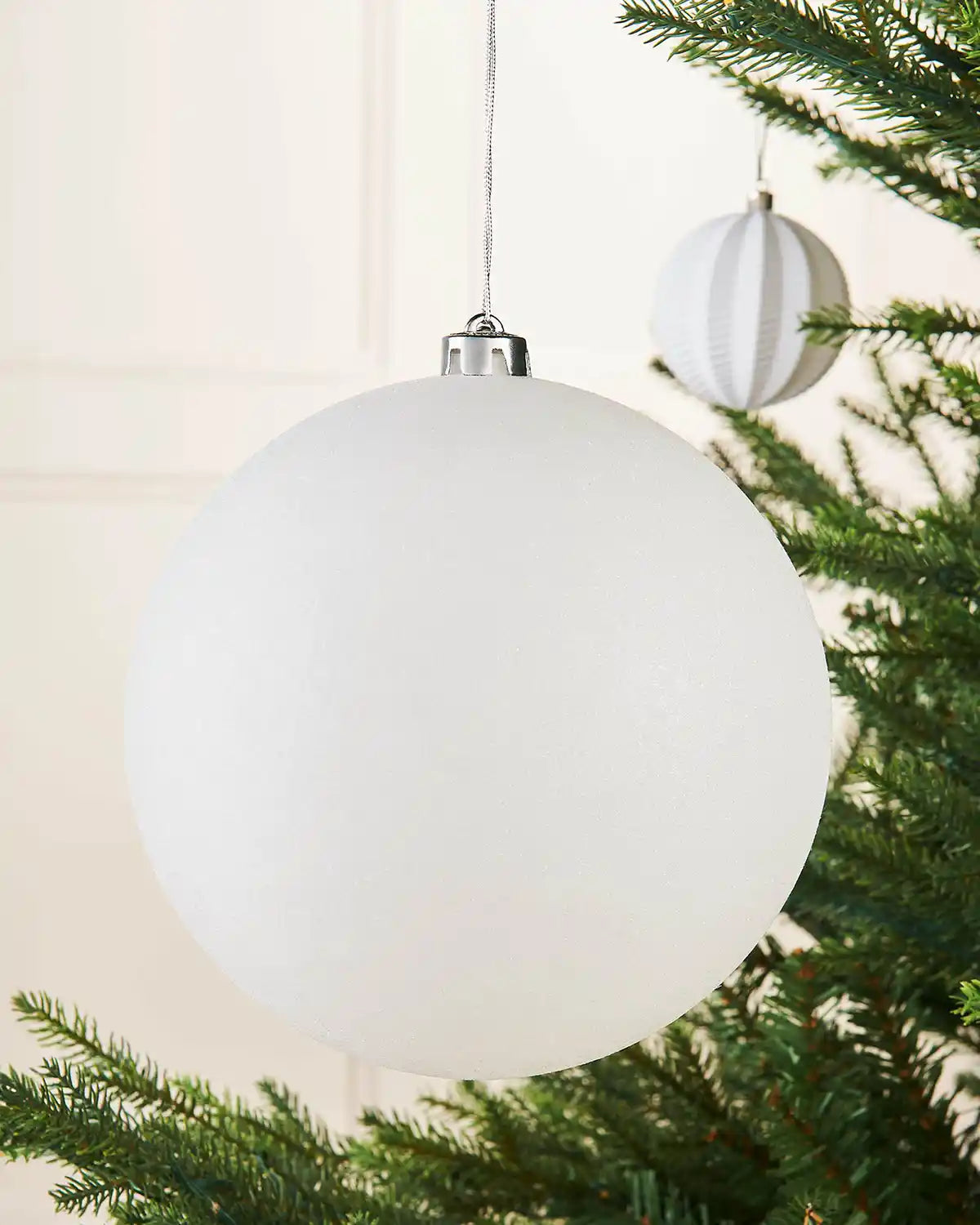 White oversized glitter bauble on a Christmas tree branch against a cream panel wall with a smaller white bauble in the background