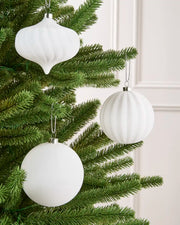 Three white ribbed Christmas tree ornaments, including two spheres and one teardrop shape, hanging from a green artificial Christmas tree against a light cream paneled wall.