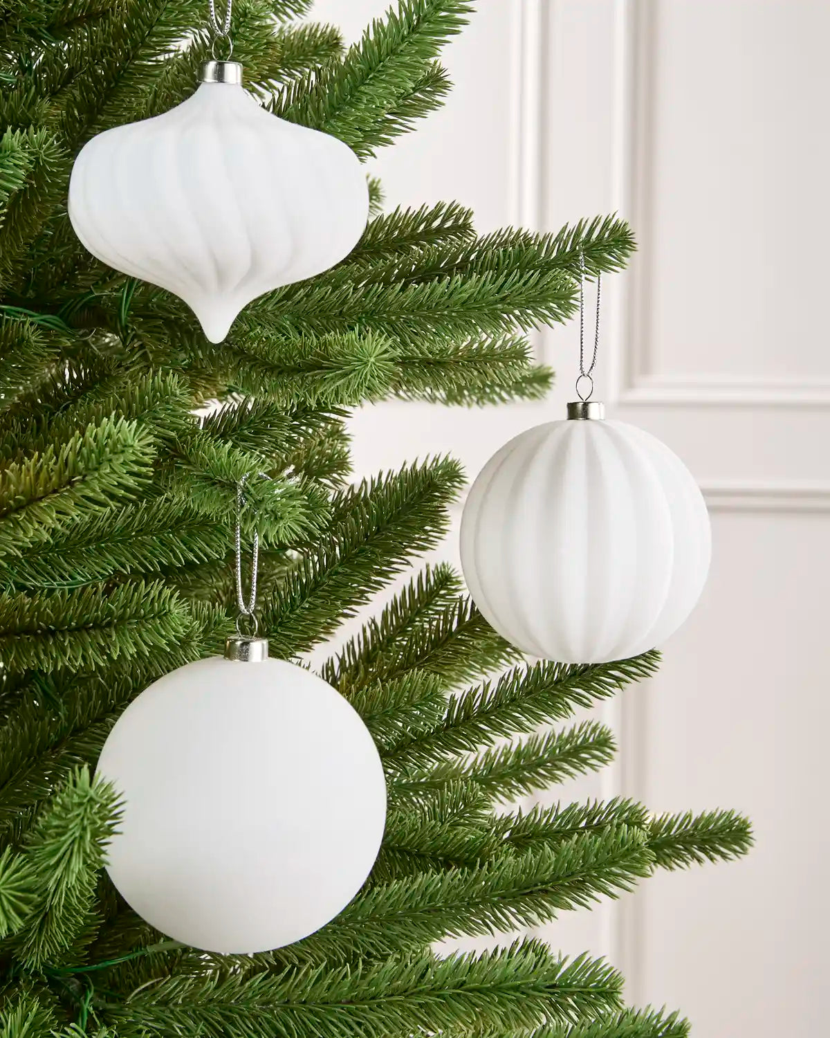 Three white ribbed Christmas tree ornaments, including two spheres and one teardrop shape, hanging from a green artificial Christmas tree against a light cream paneled wall.