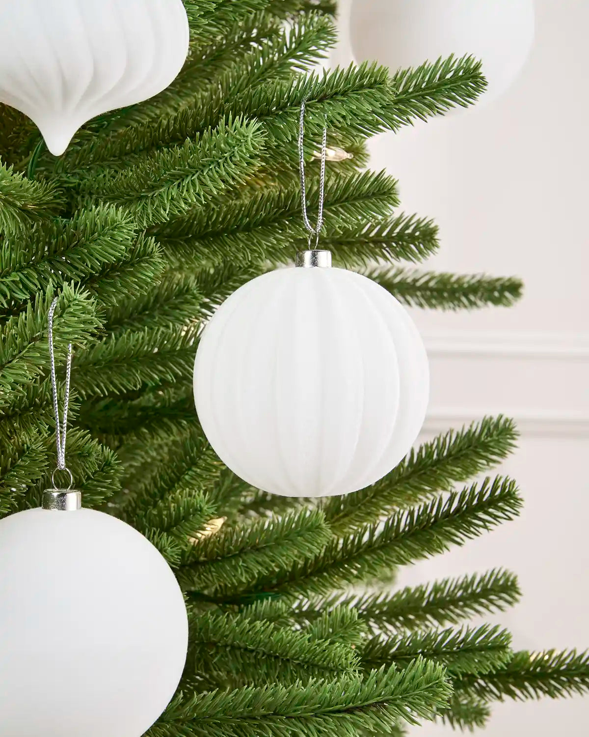 White ribbed spherical Christmas tree ornaments hanging on green artificial tree branches against a soft light background.