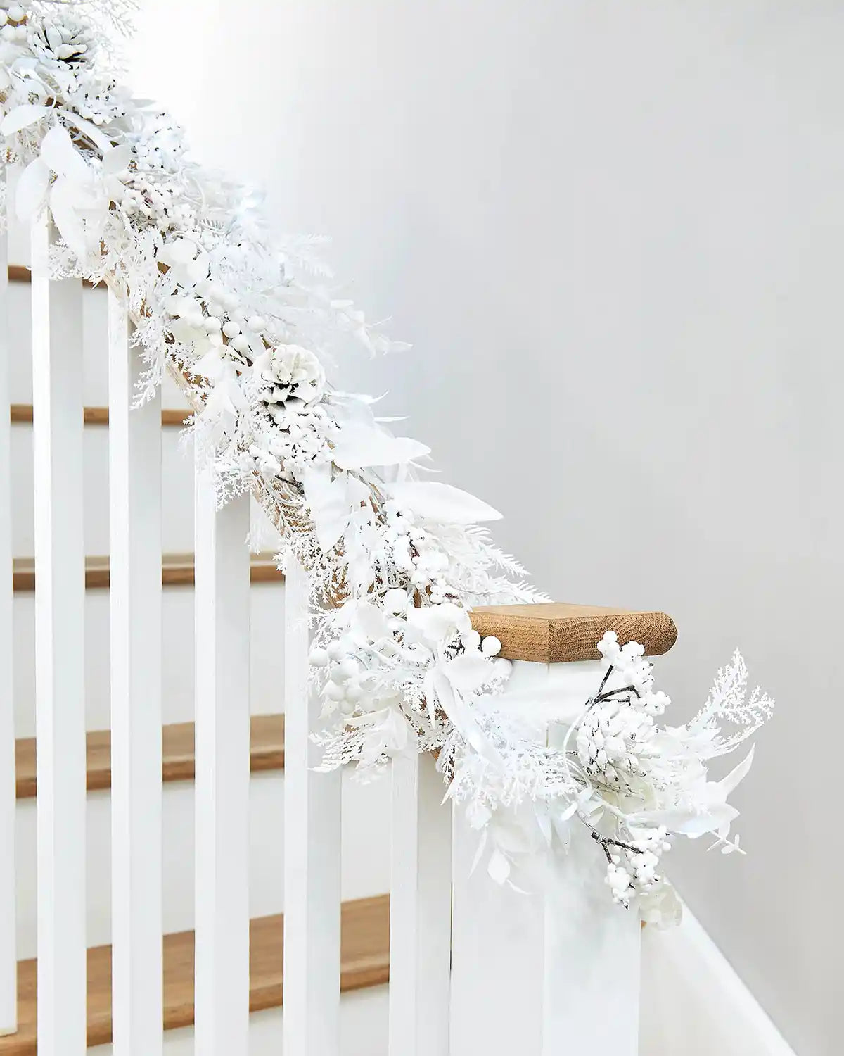 White snow flocked garland with berries and pinecones on a staircase banister against a light grey wall