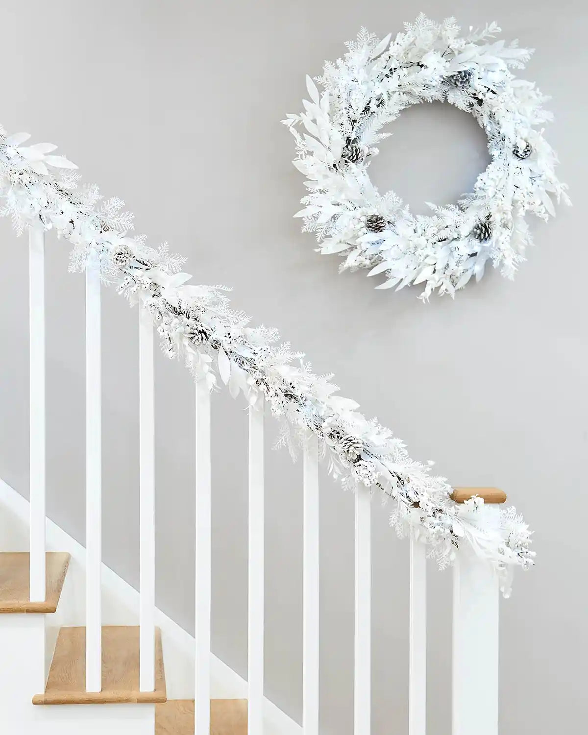 White snow flocked garland with berries and pinecones with matching wreath on a staircase banister against a cream wall