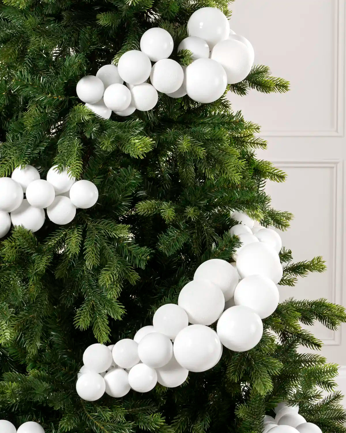 White spherical decorative ornaments draped along the branches of a green artificial Christmas tree, with a light paneled wall in the background.