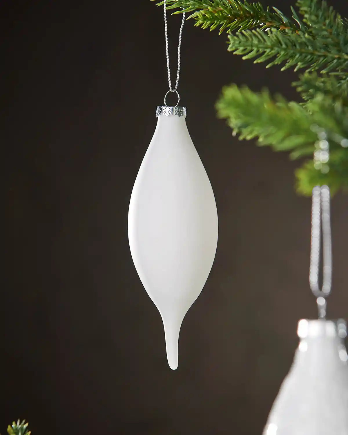 White teardrop-shaped Christmas ornament hanging on a tree branch against a dark background