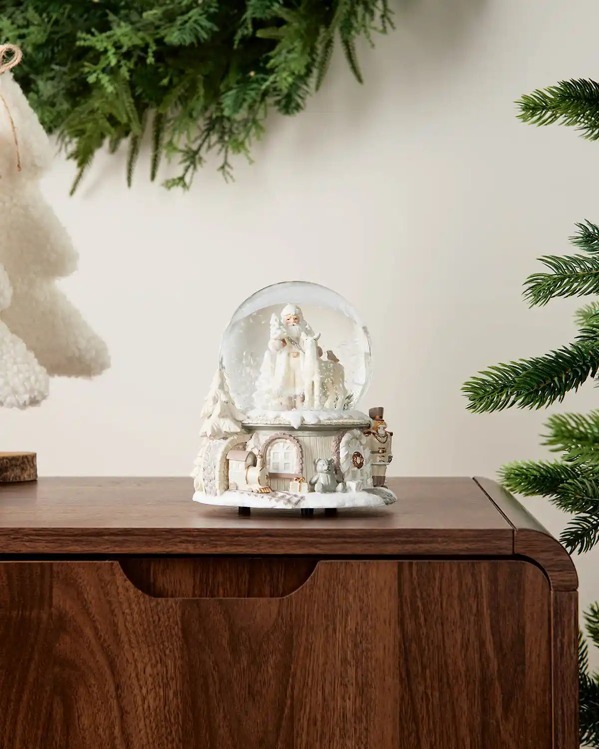 Zoomed out image of white Santa snow globe on dark wood surface in front of white wall background