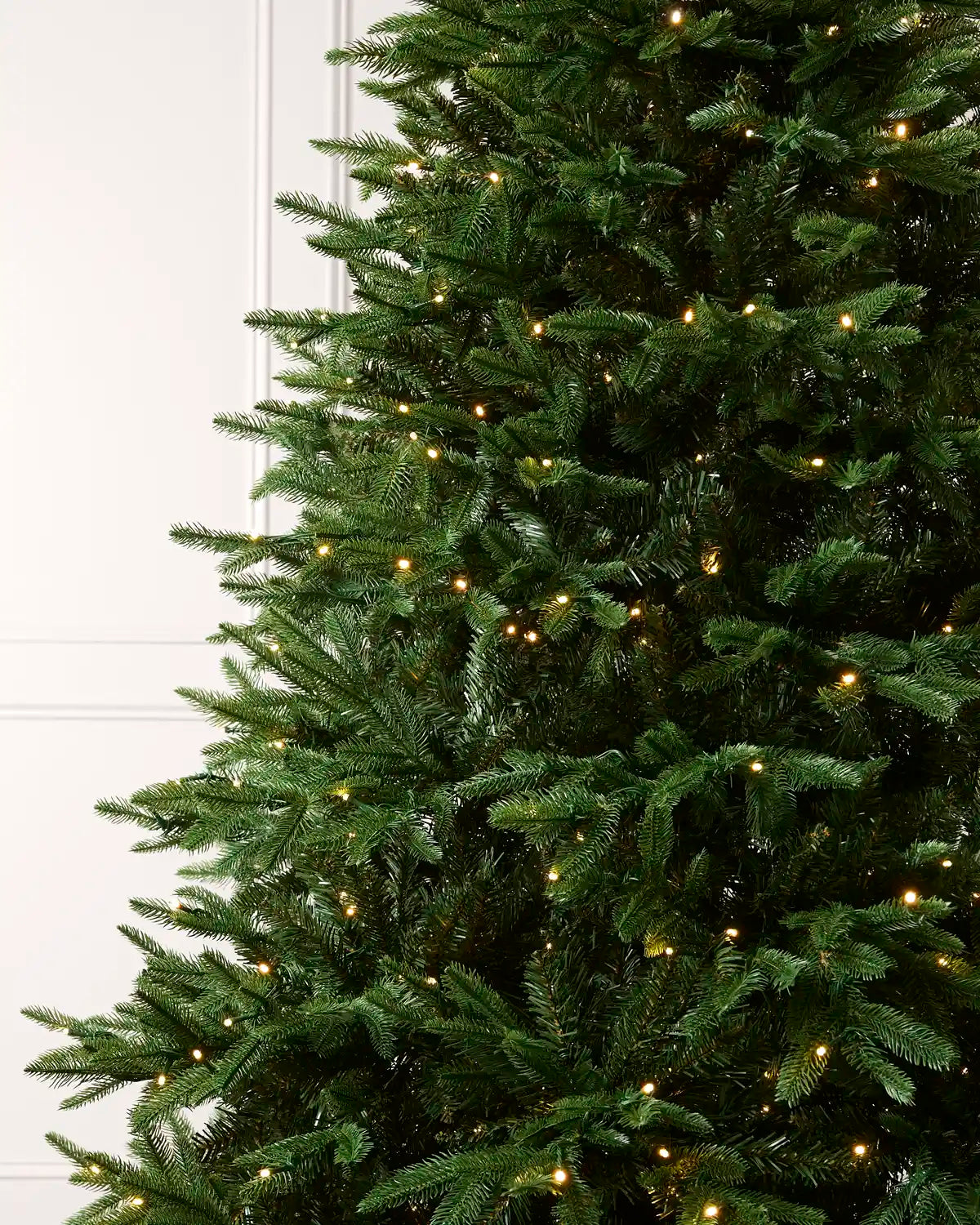 A close-up of a green artificial Christmas tree with warm white string lights, shown against a white paneled wall.