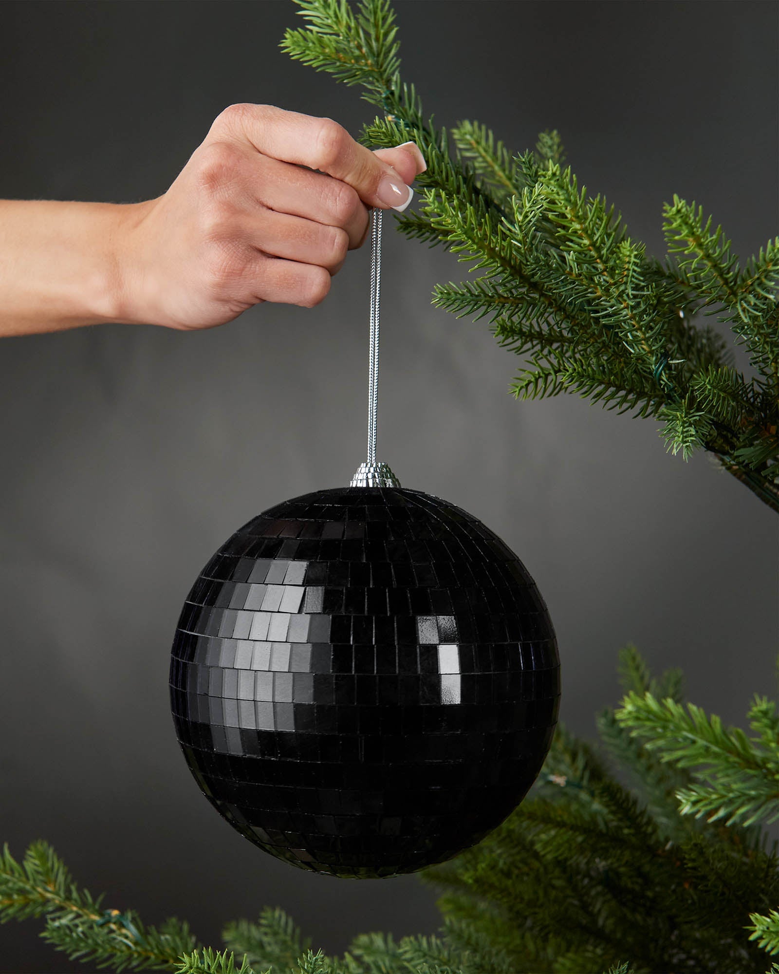 Hand holding a black 15cm oversized disco ball bauble beside a Christmas tree.