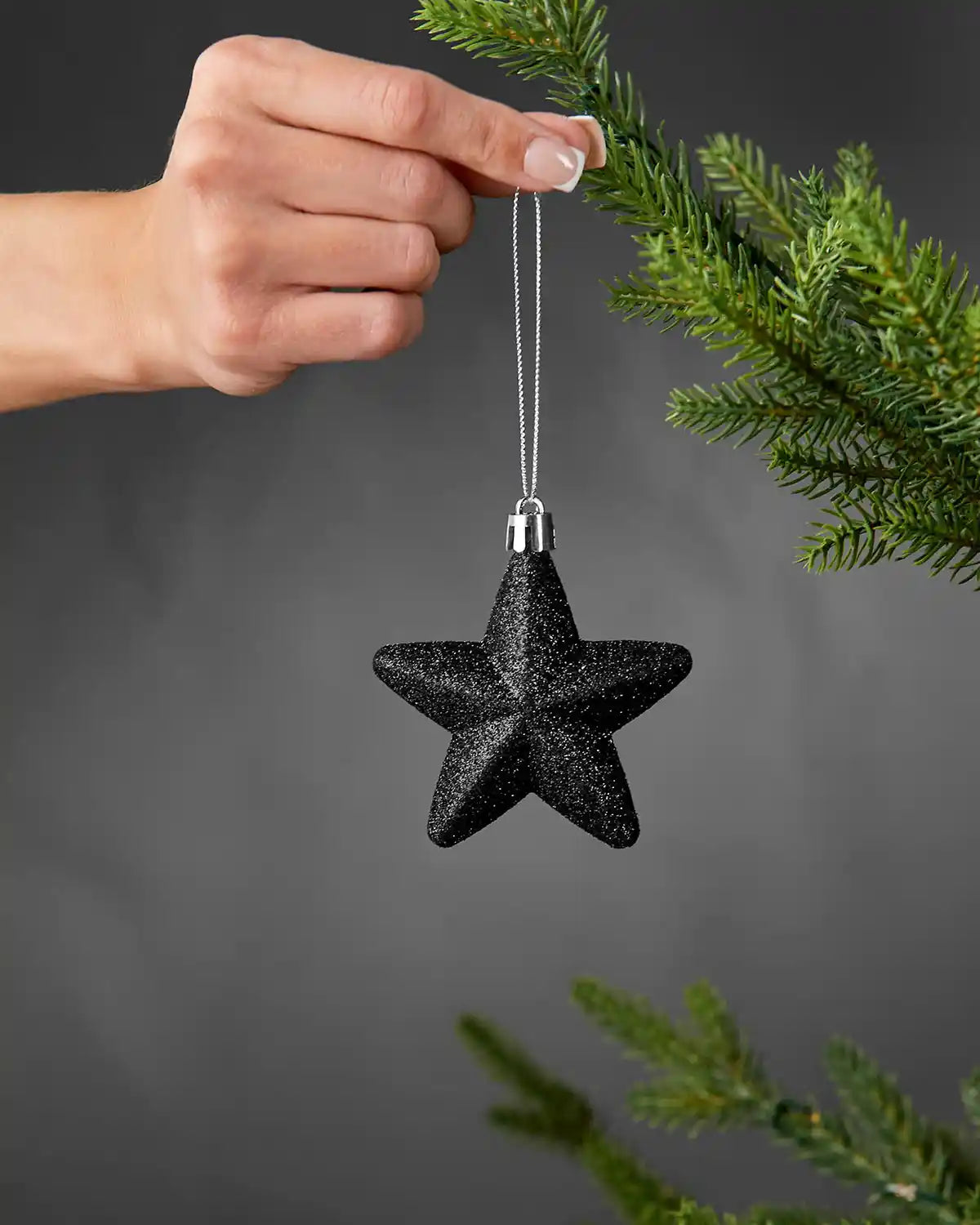 Hand holding a black 7cm glitter star decoration beside a Christmas tree.