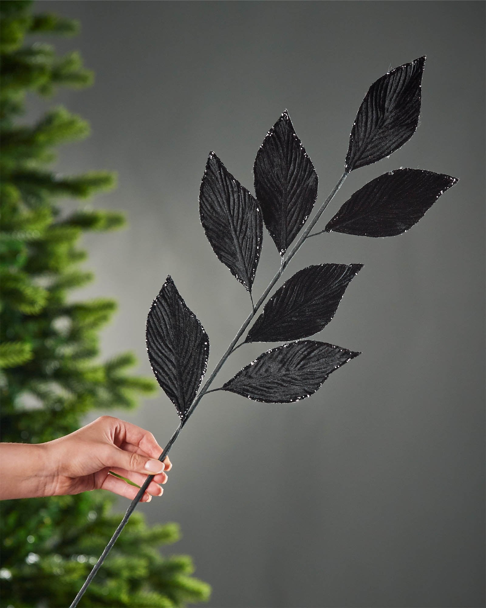 72 cm black artificial leaf spray held beside a Christmas tree, showing the full stem and elongated glitter-edged leaves.