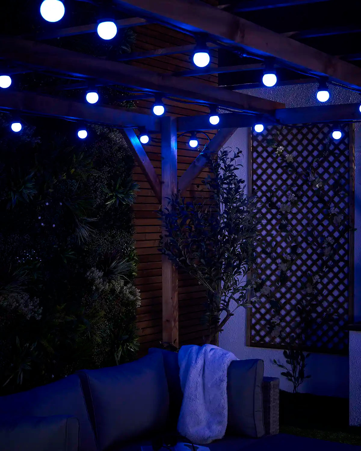 Close-up of blue E27 festoon bulbs glowing beneath a wooden pergola with outdoor seating below.