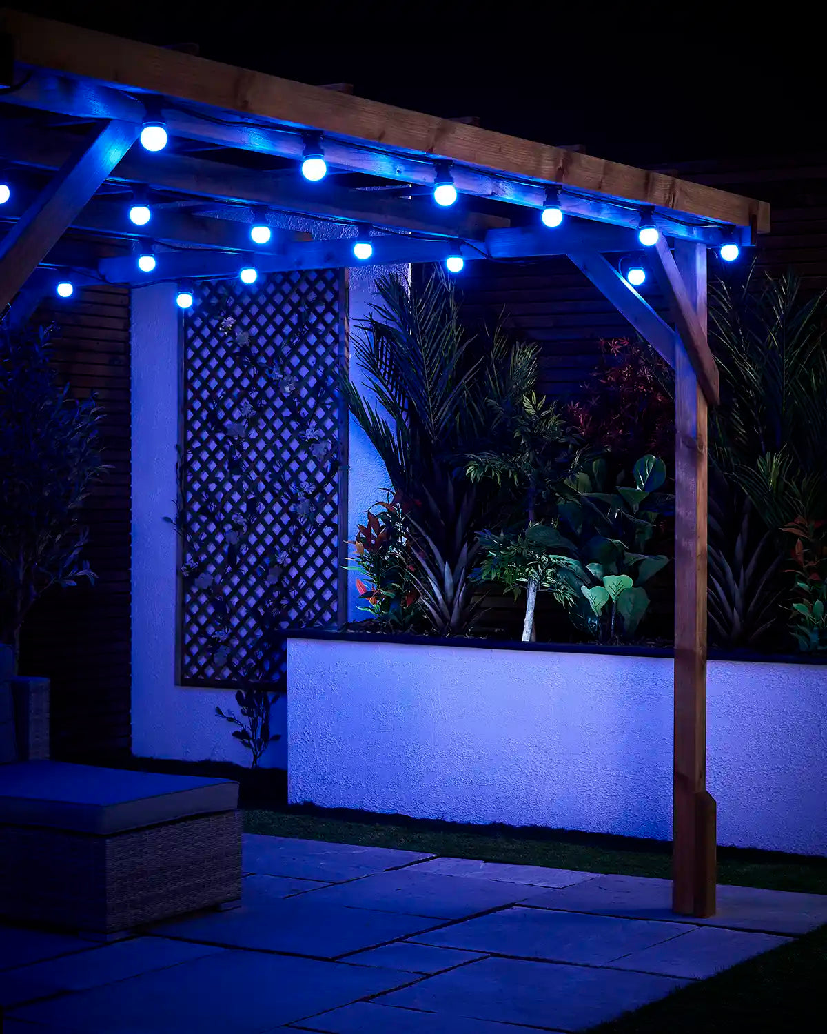 Blue E27 festoon bulbs hanging from a wooden pergola above a paved patio and raised garden bed.