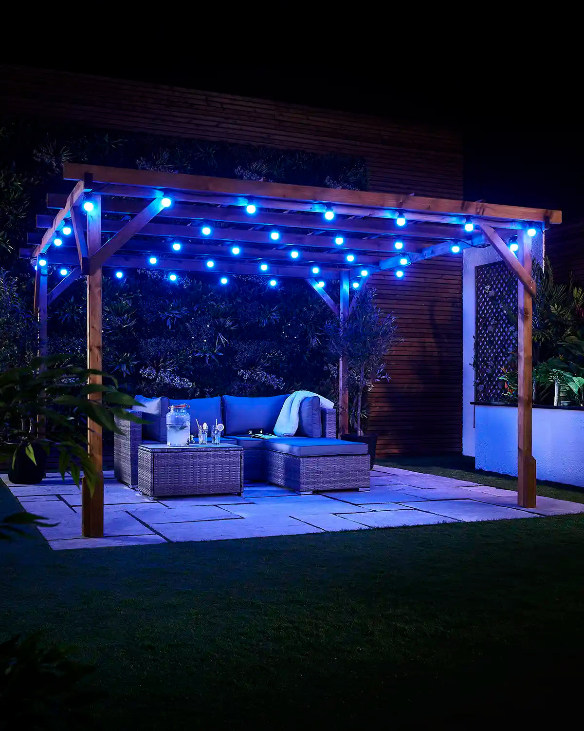 Blue E27 festoon bulbs displayed across an outdoor wooden pergola at night with patio seating and garden plants.