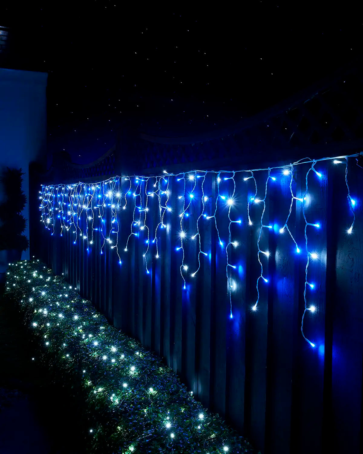 Blue and white icicle lights displayed along a garden fence above a lit hedge at night.