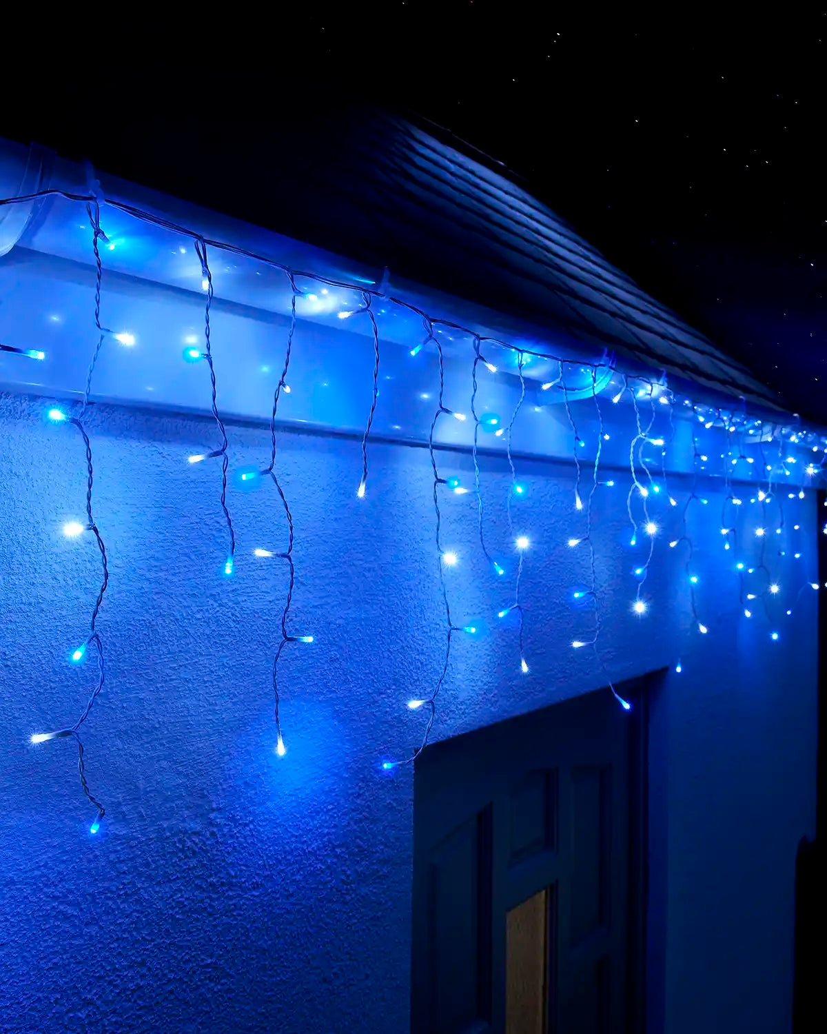 Blue and white icicle lights hanging along a roofline above an exterior wall and doorway at night.