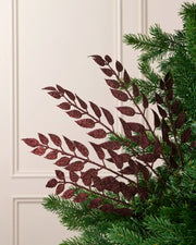 Burgundy glitter leaf Christmas tree spray arranged between green Christmas tree branches.