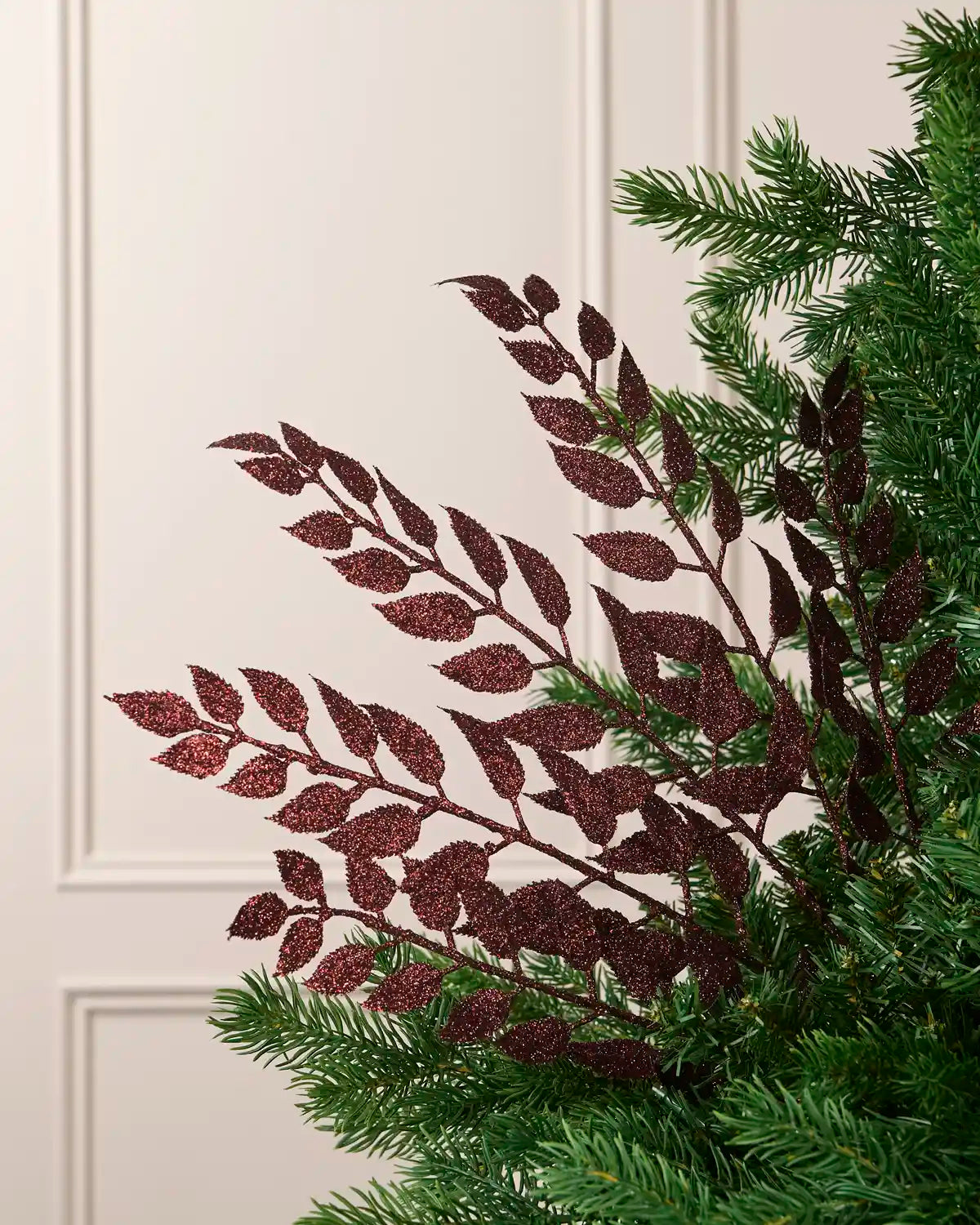 Burgundy glitter leaf Christmas tree spray arranged between green Christmas tree branches.